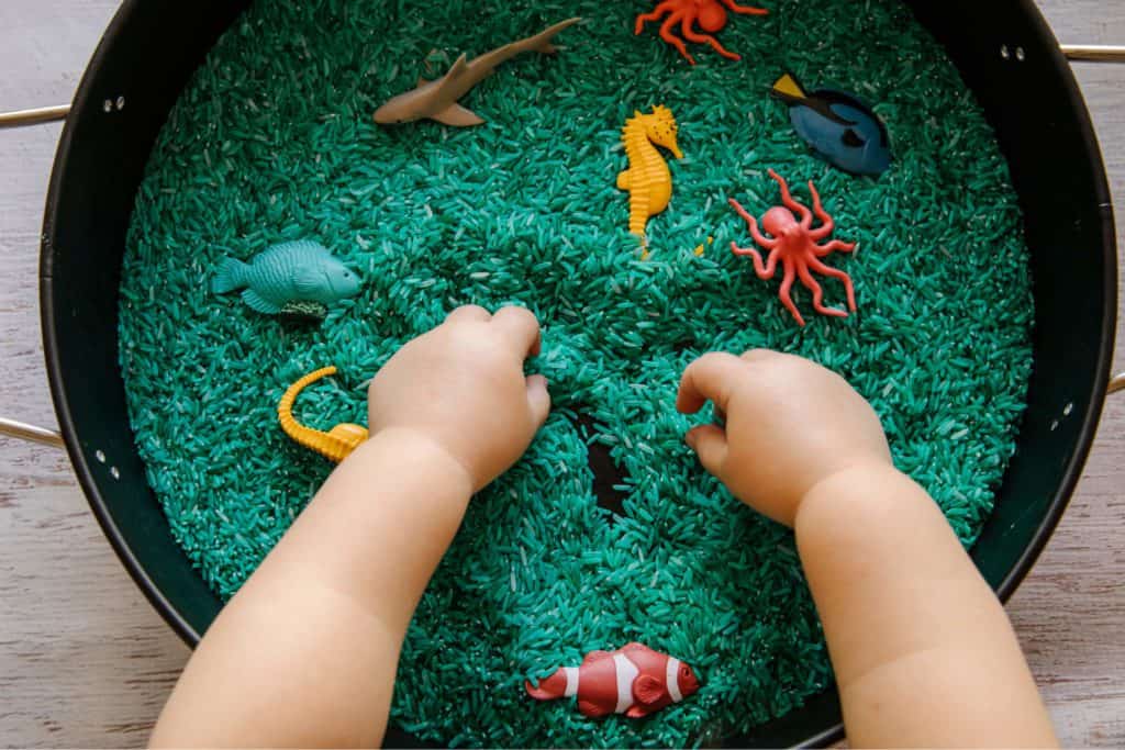 A baby/toddler enjoying a sensory bin with fish figurines and rice inside!