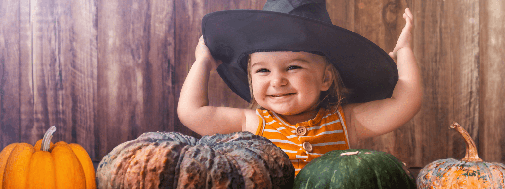 Baby giving a cheeky grin, wearing aw witches hat whilst being surrounded by pumpkins
