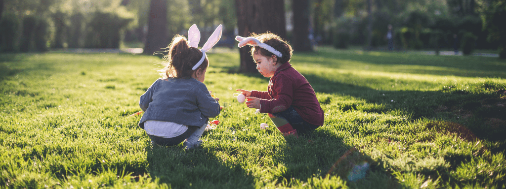 Easter Activities Toddlers: Two toddlers stand in a field holding Easter eggs and wearing bunny ears 