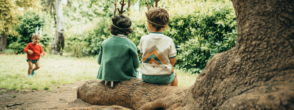 two kids sit on a tree root while a child in the background runs around. 