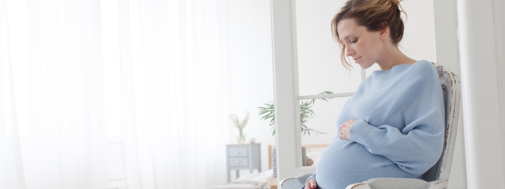 leg cramps during pregnancy -image shows a pregnant woman holding her bump while sat in a chair with a firm back.