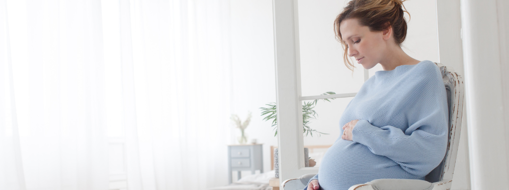 leg cramps during pregnancy -image shows a pregnant woman holding her bump while sat in a chair with a firm back. 