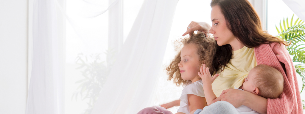 A mum, toddler and baby hug on a bed in front of a curtain