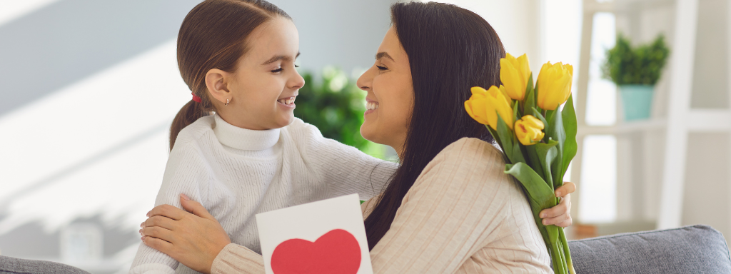 A girl gives her mum a mother's day card and a bunch of yellow flowers as they both hug and smile