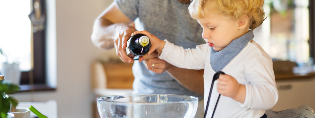 getting children involved in the ktichen -image shows a toddler (assisted by their dad) pouring dressing onto a bowl of salad